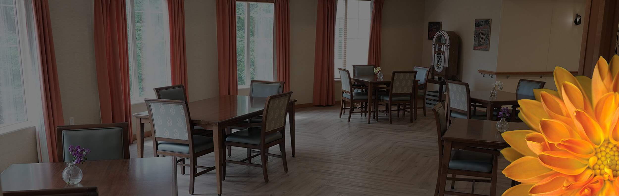 A bright and inviting dining area at The Waters of Plymouth, featuring several wooden tables with cushioned chairs, large windows with red curtains allowing natural light to flood the room.